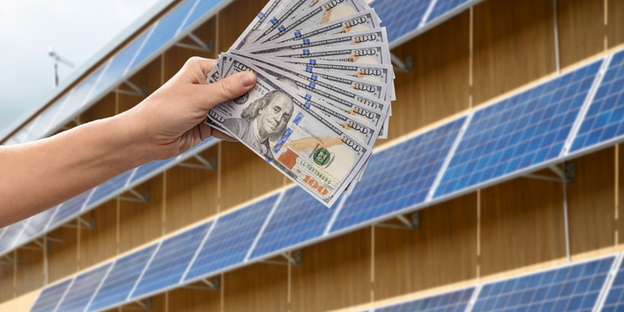 Hand houdt een waaier van bankbiljetten voor een rij zonnepanelen, symbool voor energiebesparing.